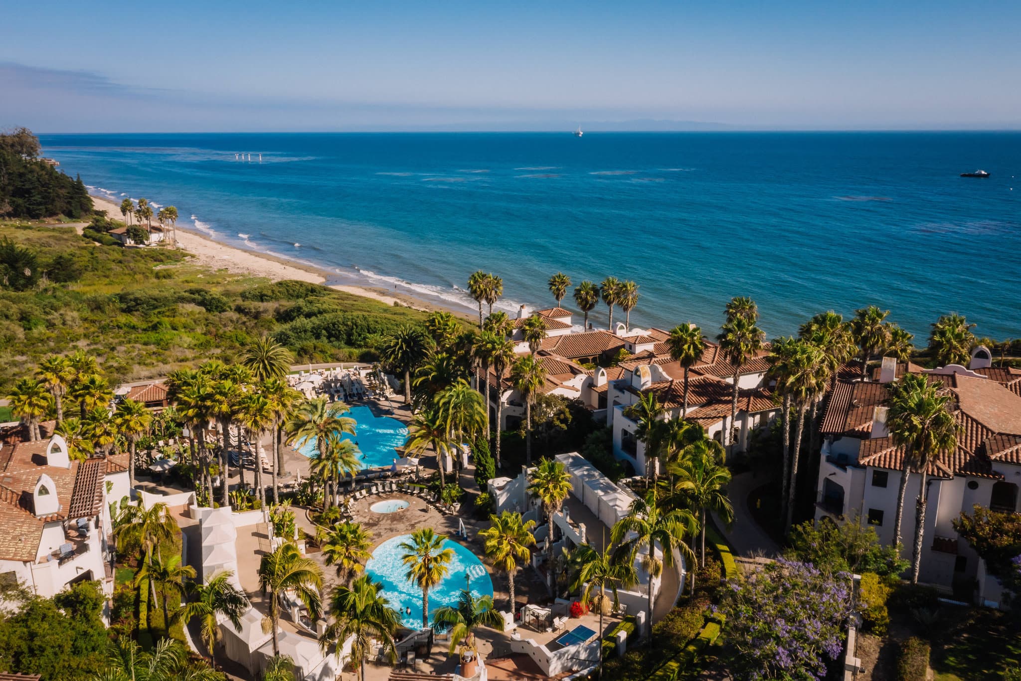 A sweeping view of The Ritz-Carlton Bacara's zero-edge pool reflecting palm trees and the Pacific Ocean at sunset, framed by Spanish-Colonial white stucco architecture.