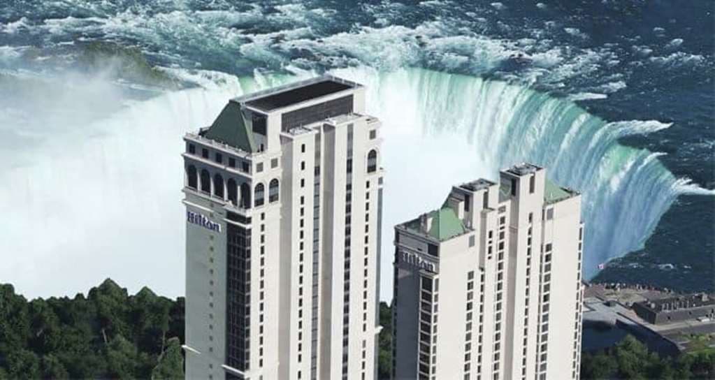 A silhouetted figure relaxing in an in-room jacuzzi tub, looking out a floor-to-ceiling window at the illuminated Horseshoe Falls at twilight.