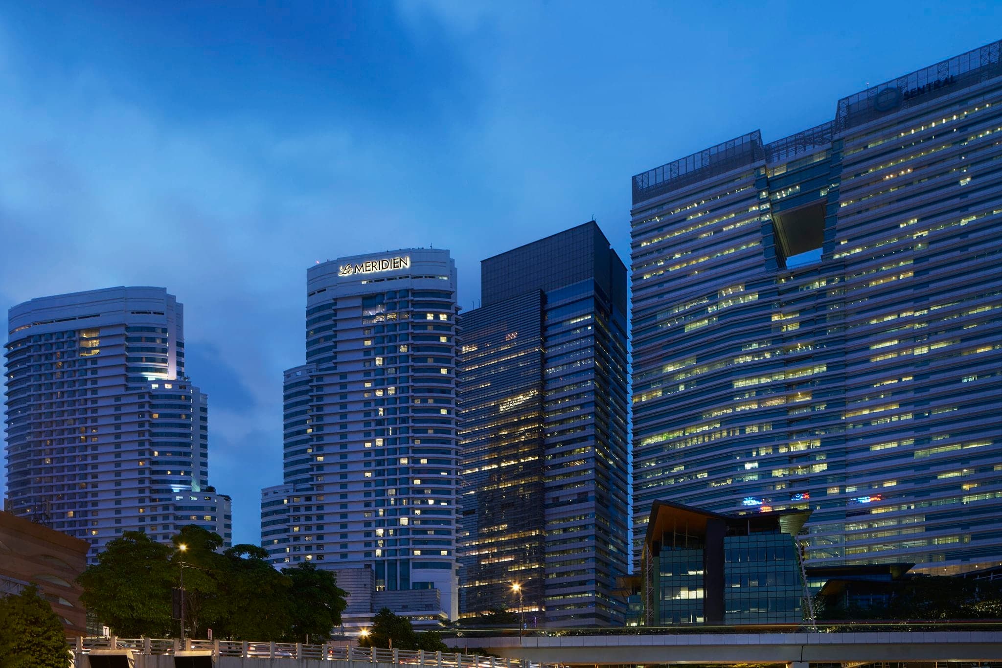A lush, tropical free-form swimming pool surrounded by palm trees and deck chairs at Le Méridien Kuala Lumpur, with the city skyline in the background.