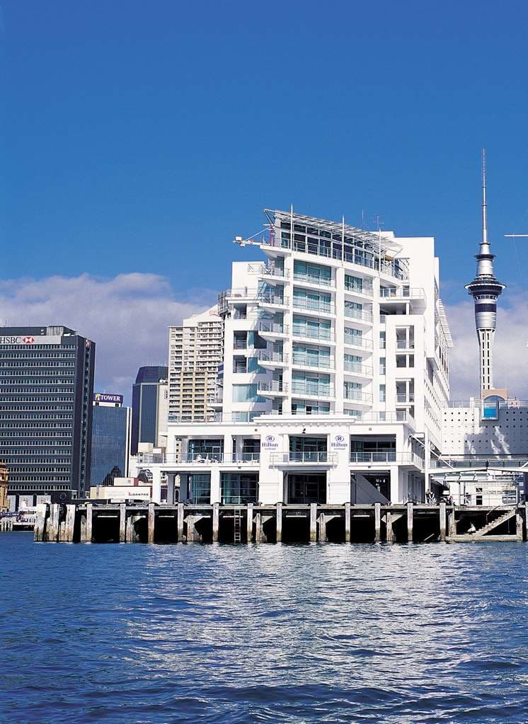 The white, ship-like exterior of Hilton Auckland extending into the harbour at sunset.