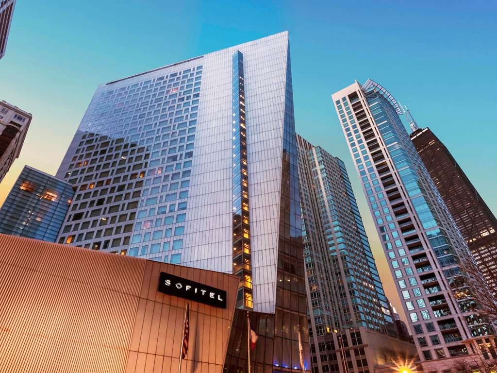 The sharp, knife-edge glass facade of Sofitel Chicago Magnificent Mile rising against the blue sky.