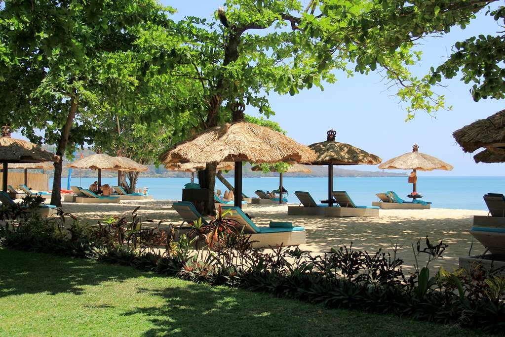 Emerald infinity pool at Jimbaran Puri bordered by stone statues and palm trees overlooking the ocean at sunset.