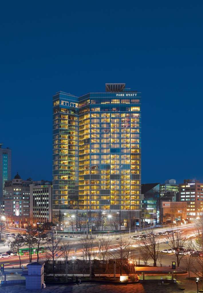 A minimalist luxury hotel room with floor-to-ceiling windows overlooking the Seoul skyline at dusk, featuring a large granite bathtub and warm wood paneling.