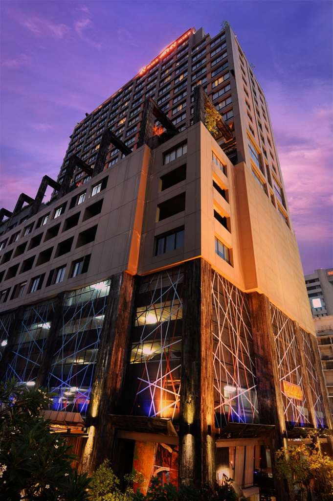 A vibrant, industrial-chic hotel lobby featuring raw concrete columns, colorful mid-century modern furniture, and floor-to-ceiling art installations.