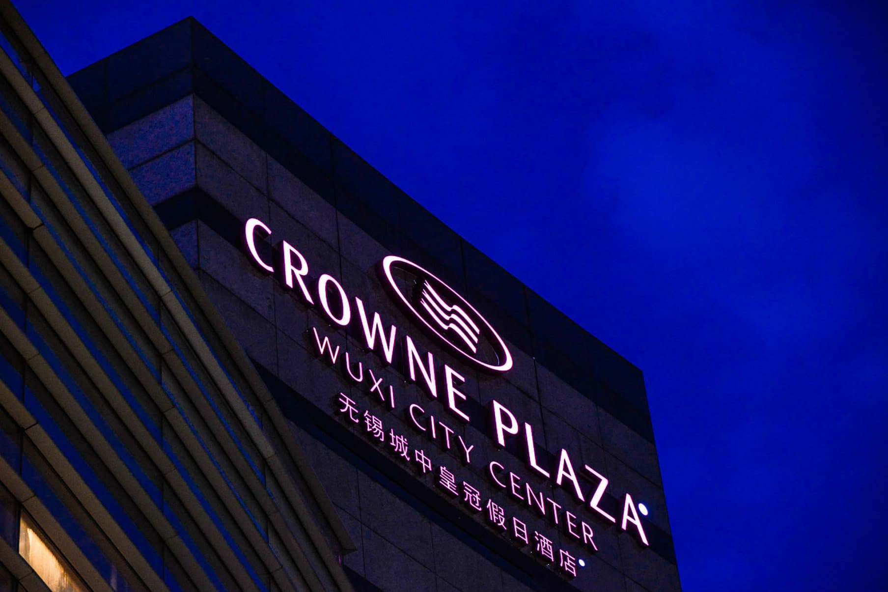 A low-angle evening shot of the Crowne Plaza Wuxi City Center's twin towers illuminated against the night sky, with the glass facade of the indoor pool glowing warm yellow.