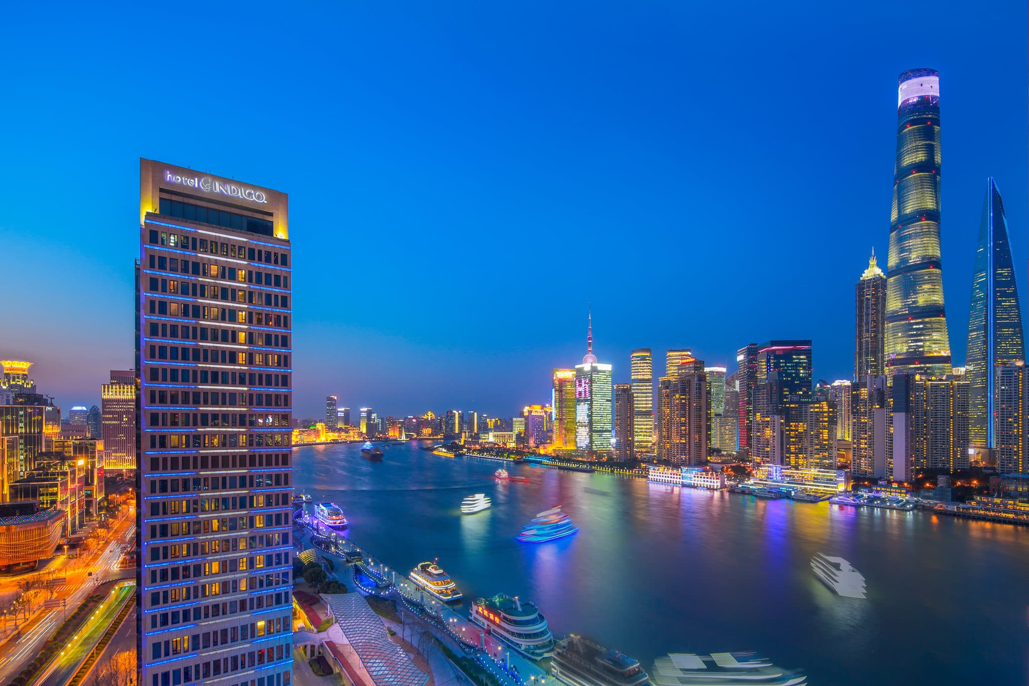 A silhouetted figure standing before a floor-to-ceiling window in a Hotel Indigo bathroom, overlooking the illuminated Shanghai Pudong skyline at dusk.