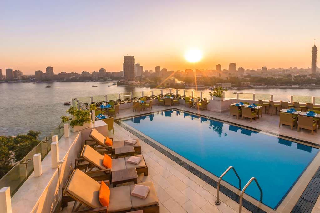 A guest balcony at the Kempinski Nile Hotel featuring a wrought-iron railing, overlooking the Nile River with felucca boats at sunset.