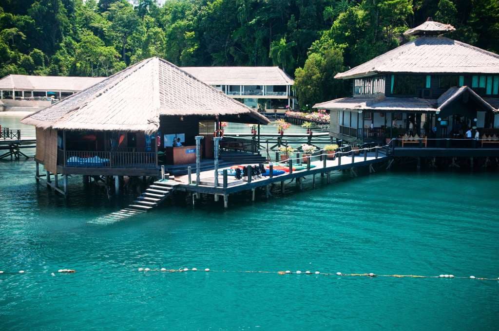 Wooden boardwalks of Gayana Marine Resort curving over turquoise water towards a lush, mist-covered rainforest mountain on Gaya Island.