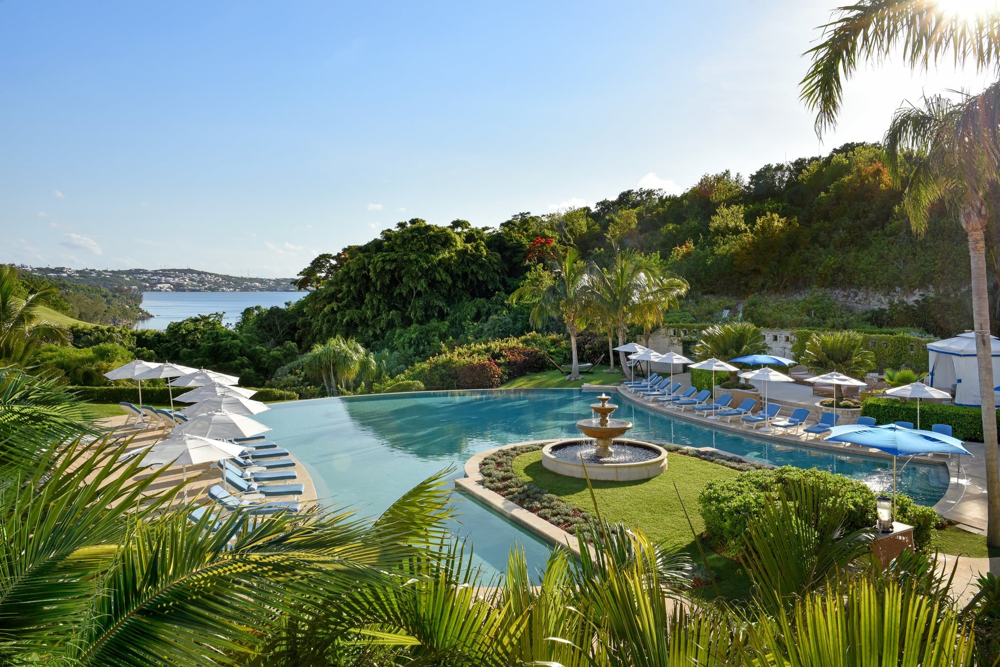 Infinity edge pool at Rosewood Bermuda overlooking Castle Harbour with palm trees and white lounge chairs.