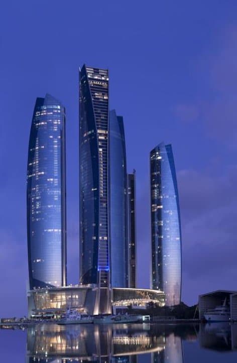 The curvaceous glass facade of Etihad Towers reflecting the Arabian Gulf sunset, juxtaposed against the white domes of the Presidential Palace.