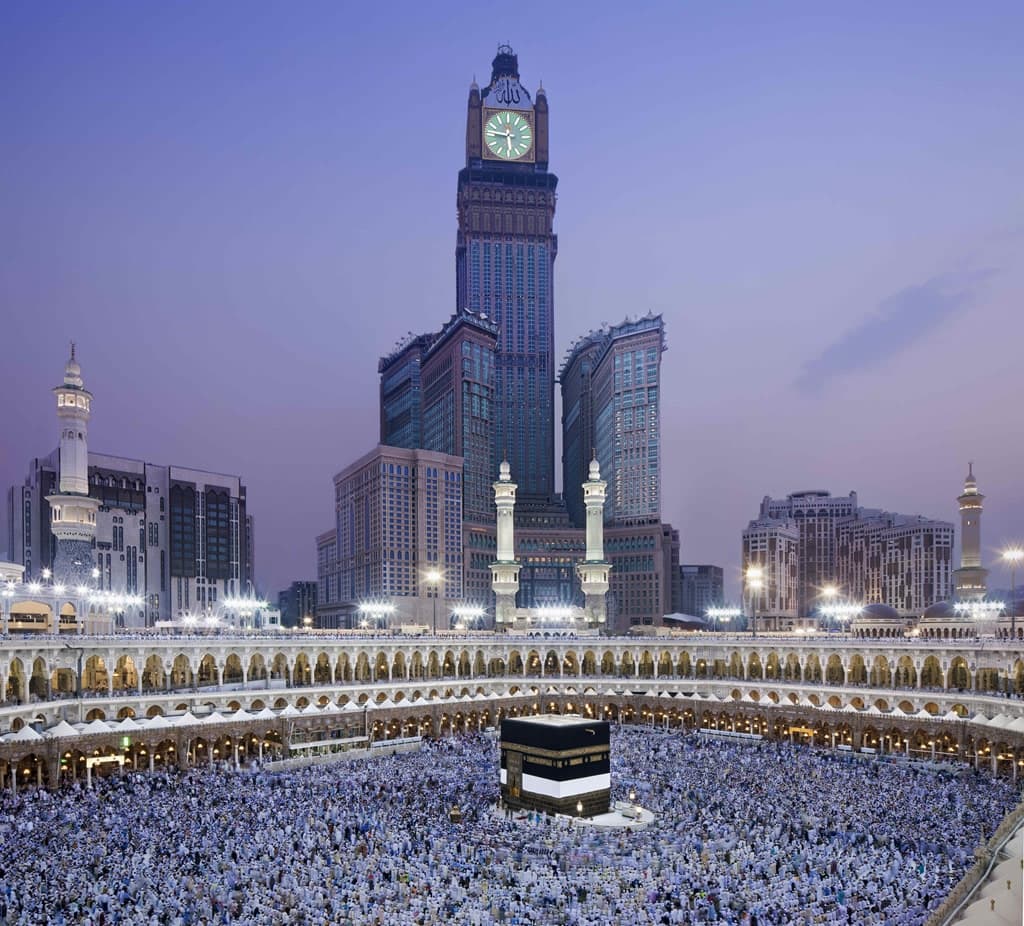A silhouette of a guest standing before a floor-to-ceiling window in a Raffles suite, overlooking the illuminated Kaaba and Grand Mosque at twilight.