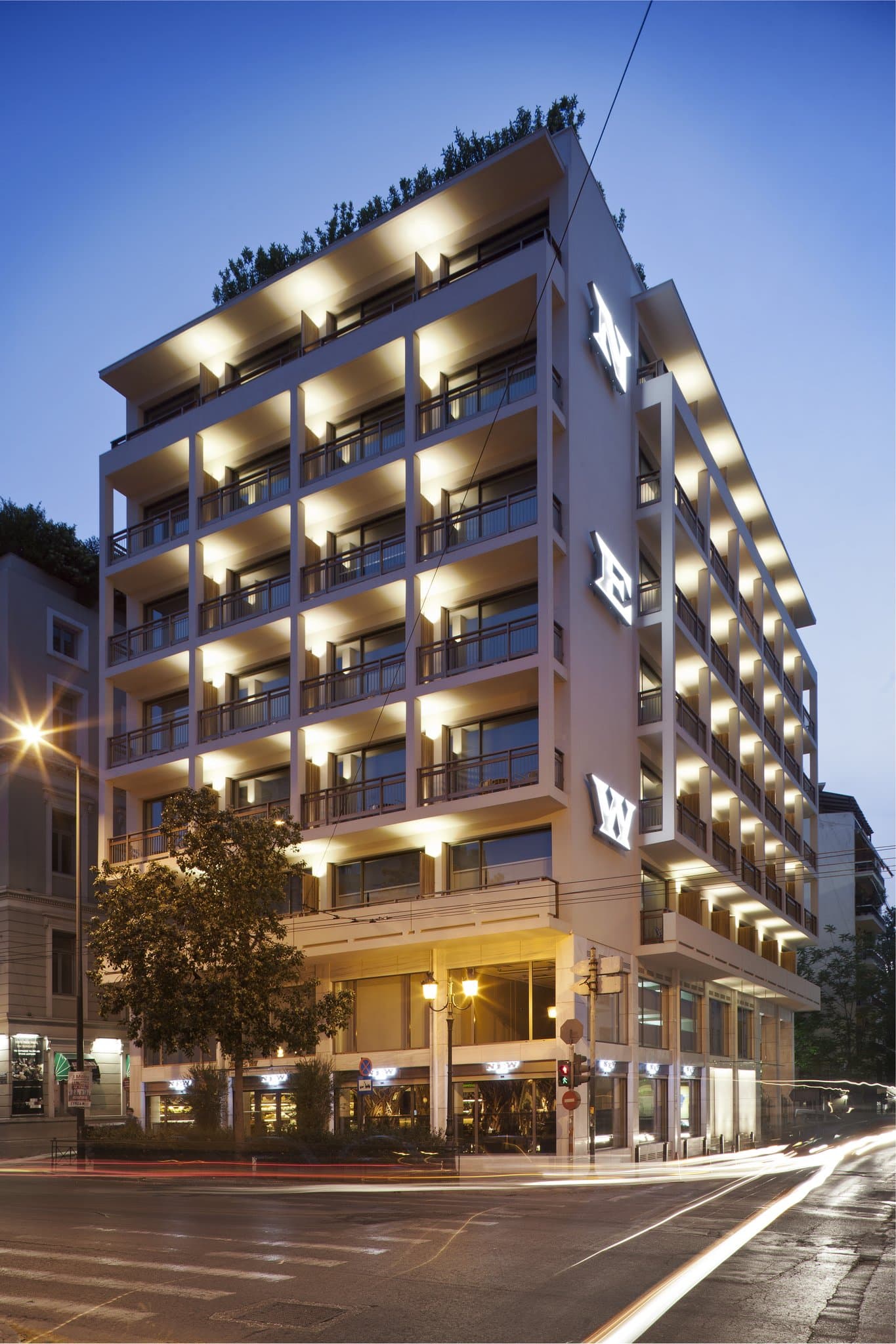 The Art Lounge rooftop at New Hotel Athens featuring a jagged library wall, modern furniture, and a direct view of the illuminated Parthenon at night.