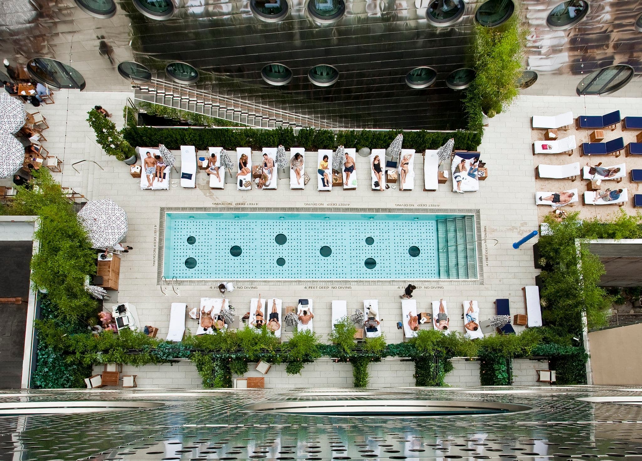 Low-angle view of the Dream Downtown lobby ceiling featuring a glass-bottom pool with swimmers visible from below, surrounded by metallic porthole-style architecture.