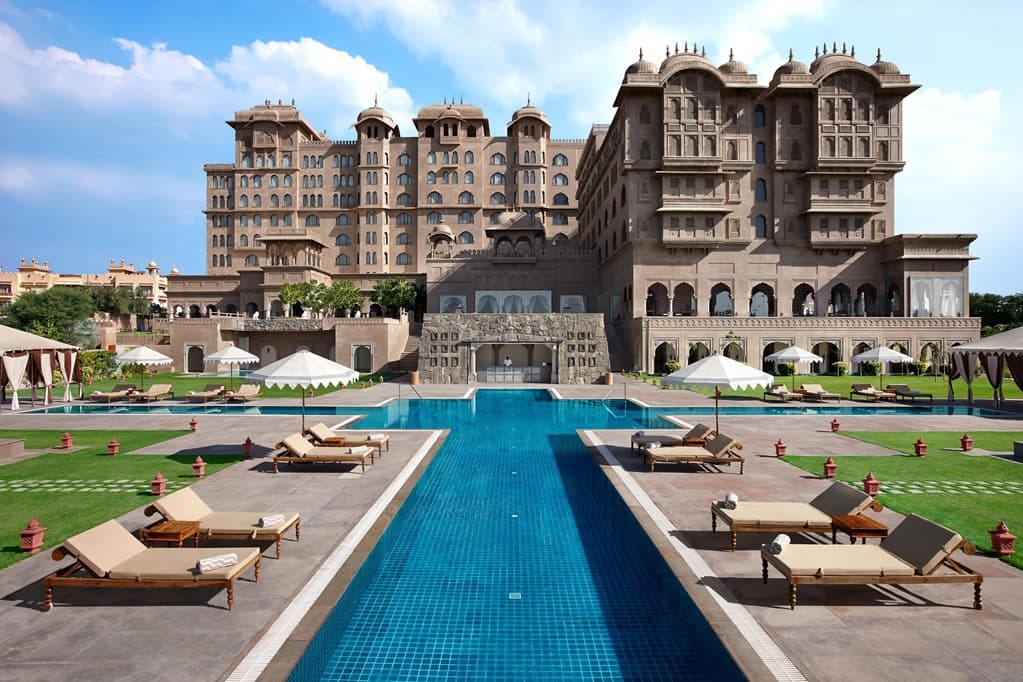 A high-angle shot of the Fairmont Jaipur's Chaupar-shaped pool, surrounded by cream-colored fortress walls and the Aravalli hills in the background.