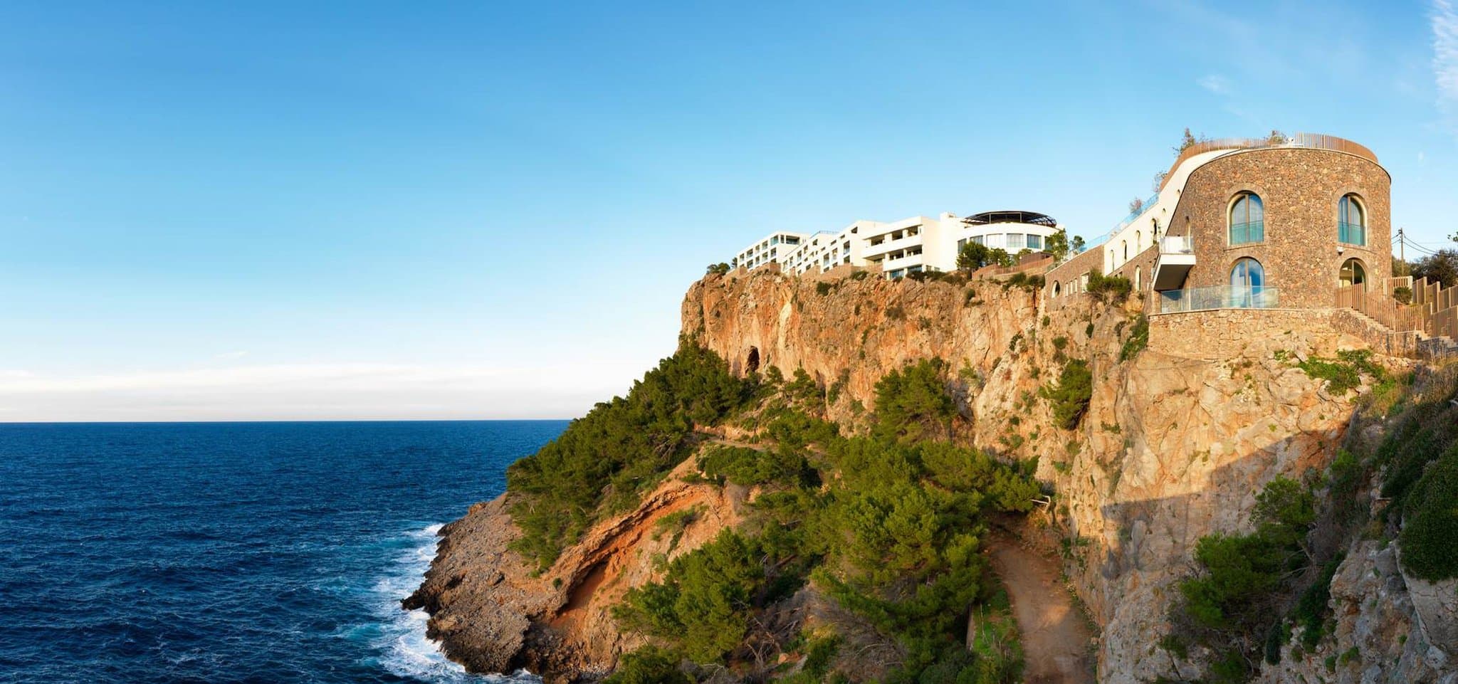 A dramatic high-angle shot of the Jumeirah Port Soller infinity pool edge blending seamlessly with the deep blue Mediterranean Sea, framed by rugged limestone cliffs.