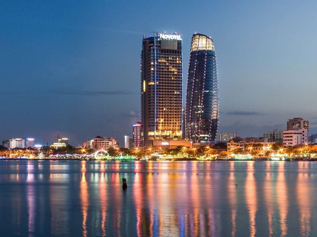 Novotel Danang Premier Han River exterior at twilight, reflecting over the water with the Dragon Bridge glowing in the background.