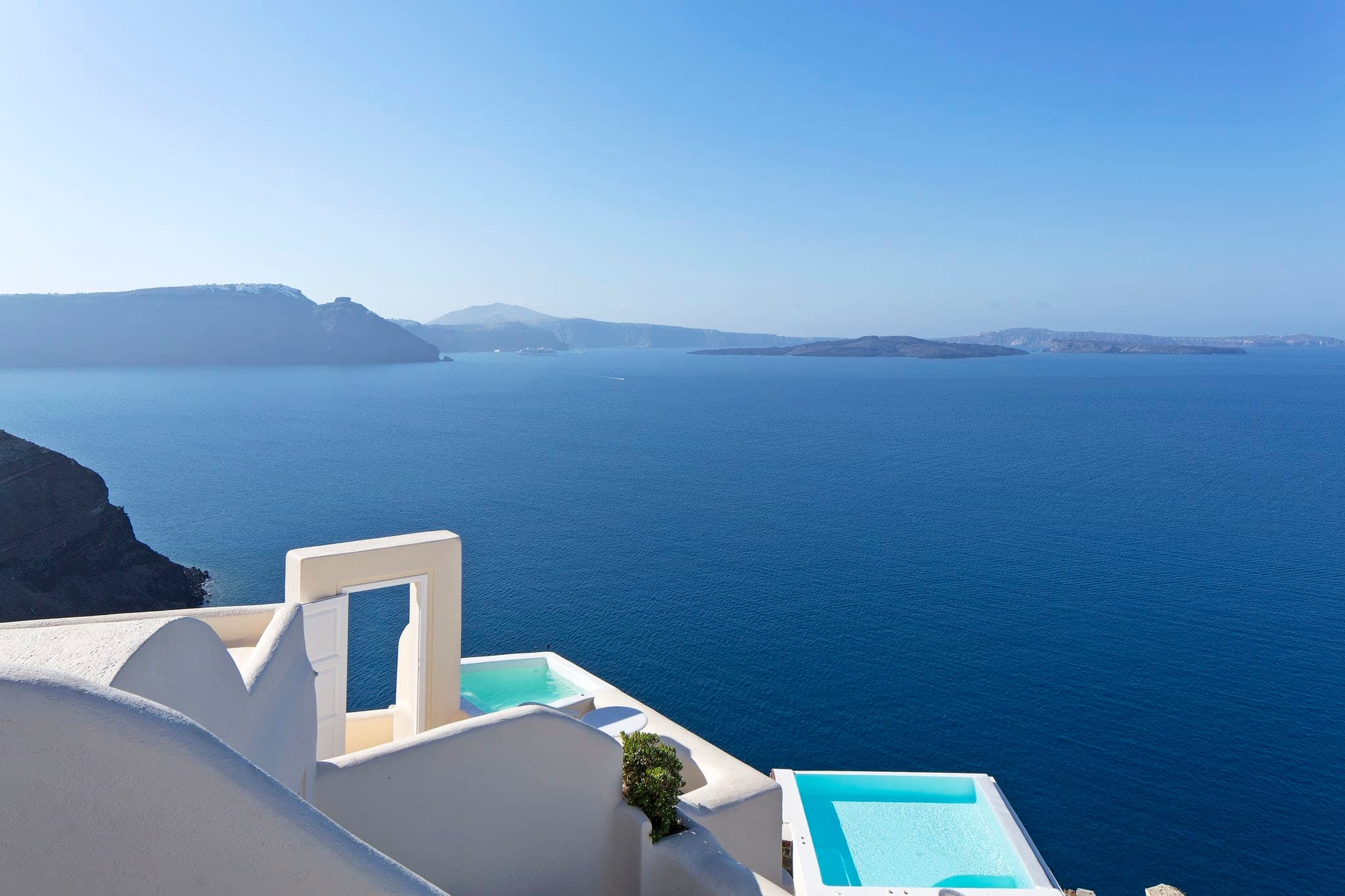 A blindingly white infinity pool at Canaves Oia Suites with floating sun loungers, overlooking the deep blue Santorini caldera.