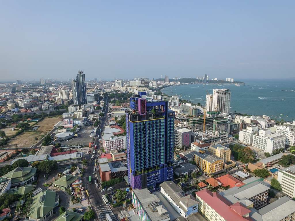 A dramatic double-decker infinity pool at sunset overlooking the Pattaya coastline, featuring guests sipping cocktails and industrial-chic metal sculptures.