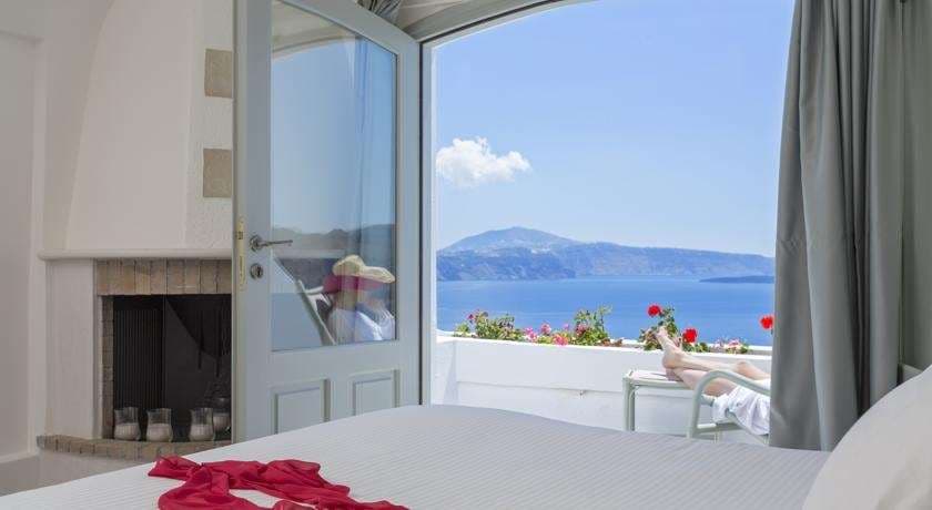 A woman in a white dress standing on a private terrace at Andronis Boutique Hotel, overlooking the deep blue Santorini caldera at sunset.