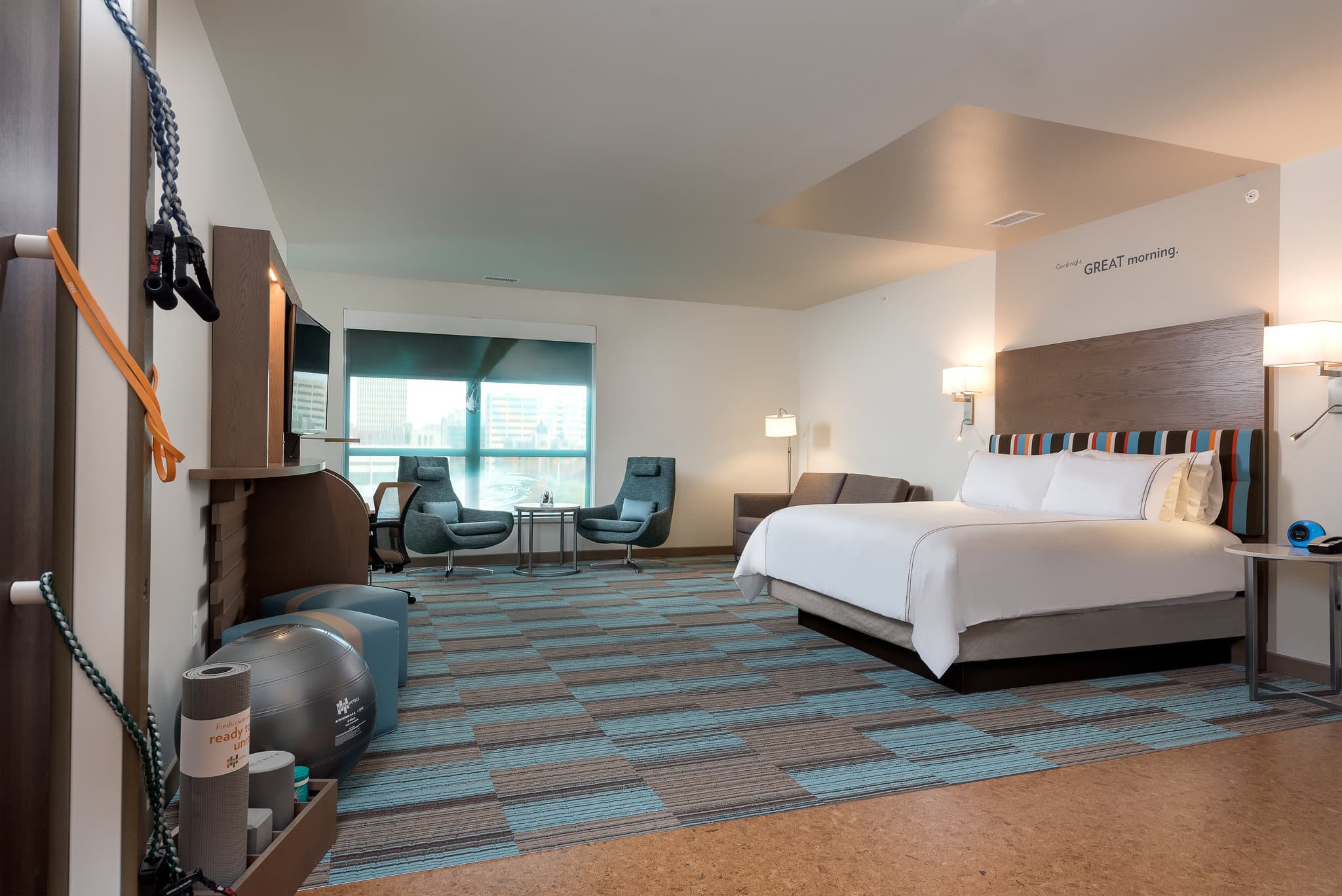 A sleek, modern hotel room featuring cork flooring, a dedicated workout corner with exercise balls and TRX straps, and a large mirror reflecting a bright, airy space.