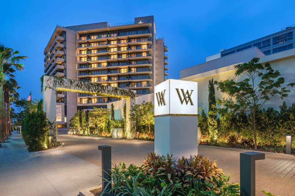 A sun-drenched rooftop pool deck at Waldorf Astoria Beverly Hills lined with private green cabanas, overlooking the panoramic Los Angeles skyline and Golden Triangle.