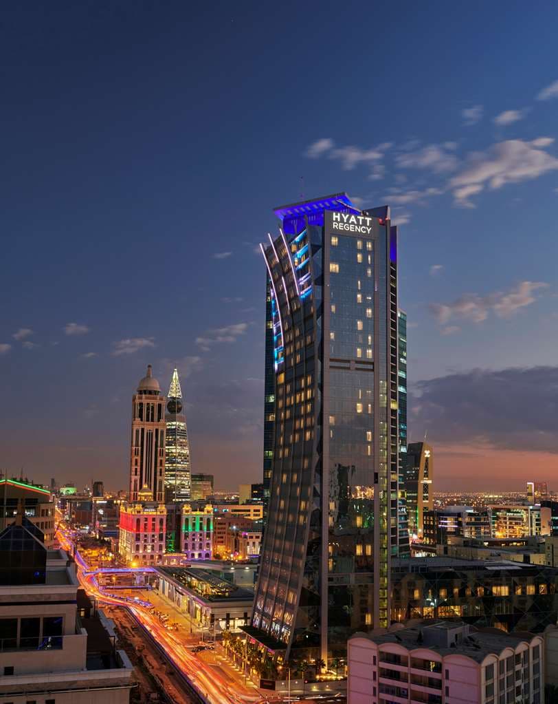 A low-angle shot of the Hyatt Regency Riyadh Olaya's glass façade reflecting the blue desert sky, contrasted against the bustling Olaya Street traffic.