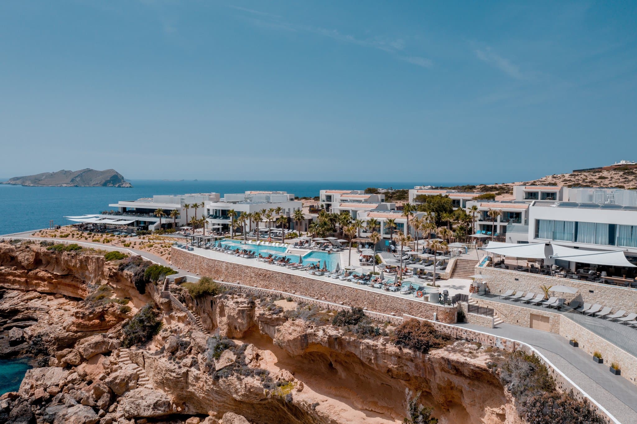 7Pines Resort Ibiza 的玻璃幕墙无边泳池,背景是夕阳下的 Es Vedrà 岩岛。