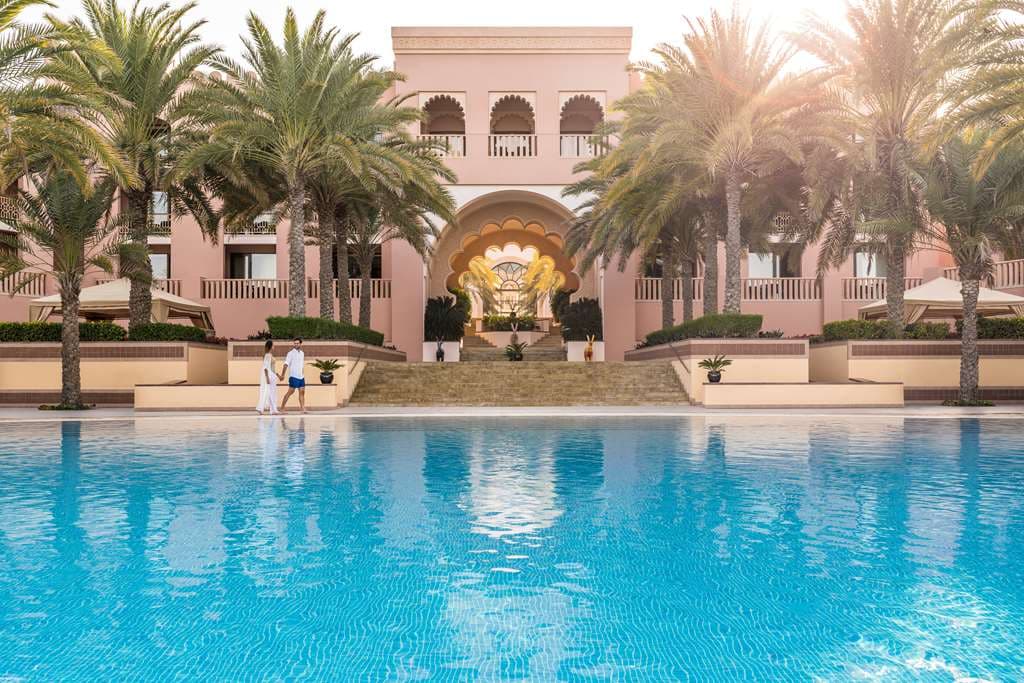 A dramatic stone archway at Al Husn Hotel Muscat framing a turquoise infinity pool and the Gulf of Oman, flanked by palm trees.