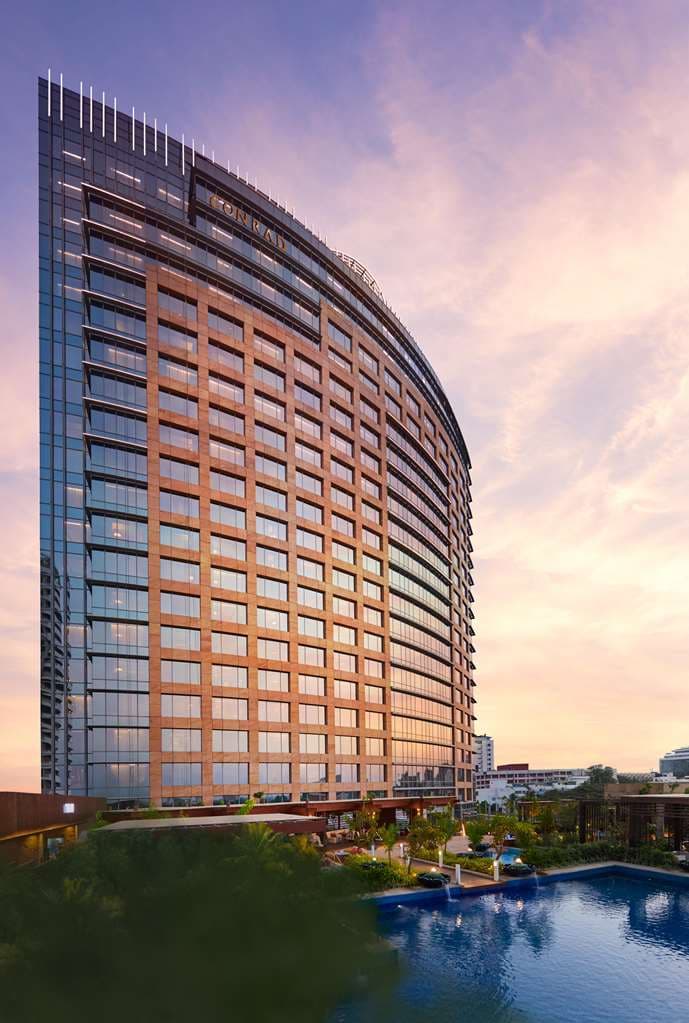 View of the curved glass facade of Conrad Bengaluru reflecting the sunset over Ulsoor Lake.