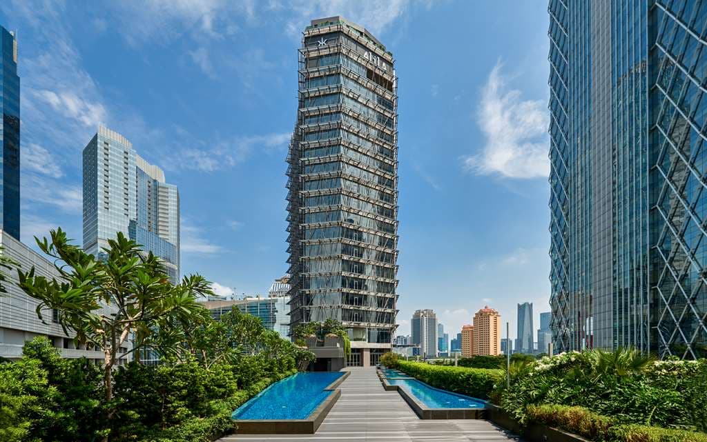 雅加达 Discovery SCBD 的 Catwalk Pool 黄昏景色,长方形泳池两侧排列着躺椅,背景是灯火通明的 SCBD 天际线和摩天大楼。