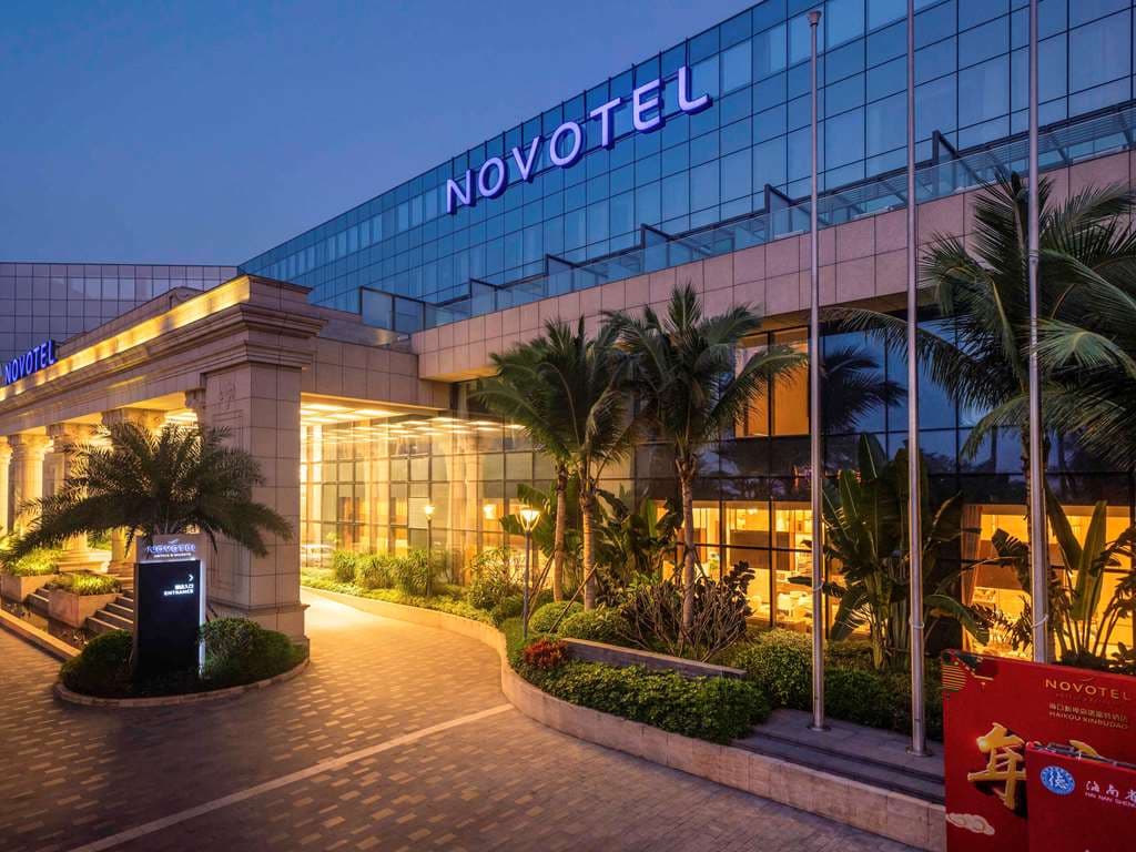 A sleek, rectangular outdoor swimming pool in the central courtyard of Novotel Haikou Xinbudao, surrounded by palm trees and modern white balconies.