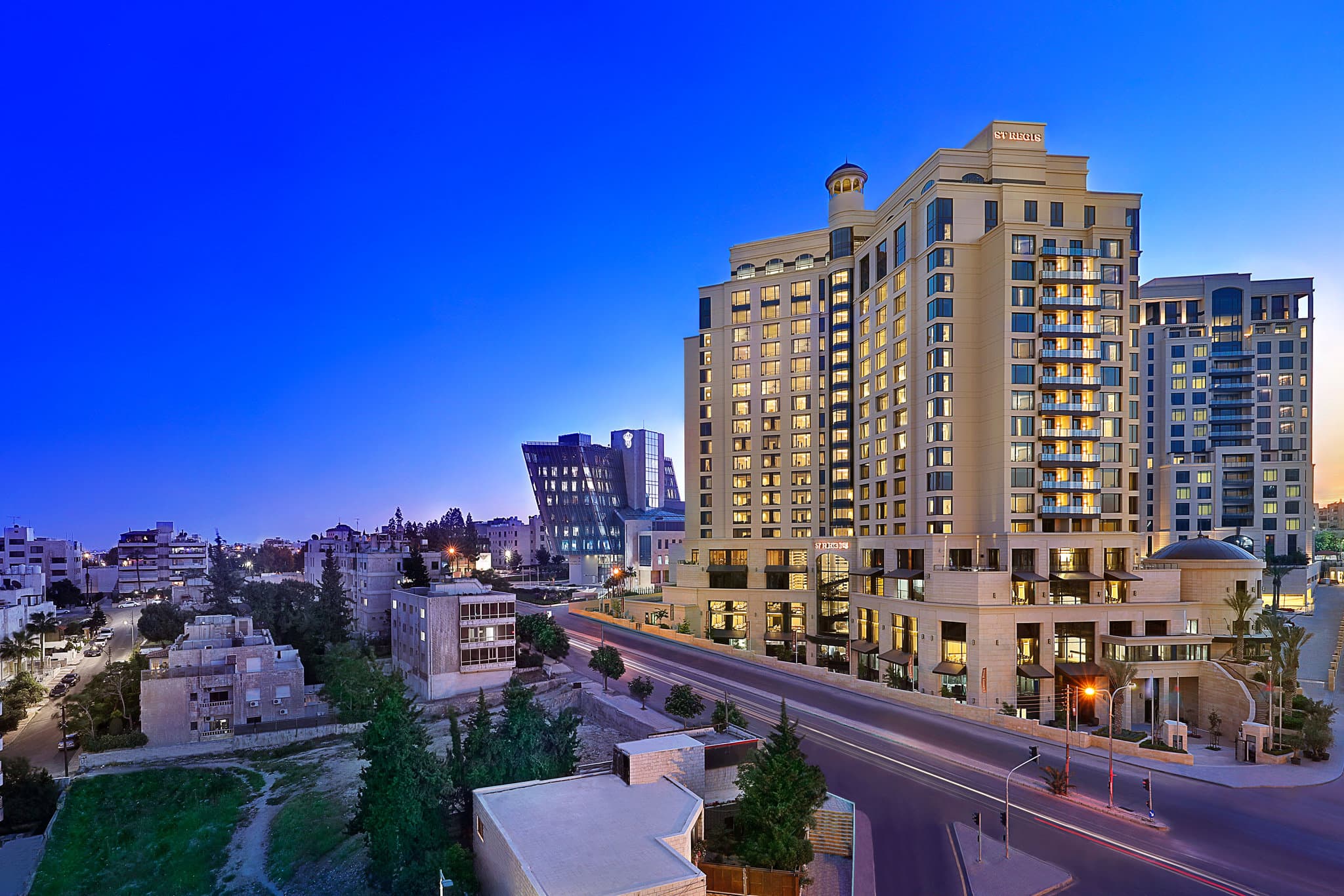 The Zenith rooftop terrace at The St. Regis Amman at night, featuring illuminated tables, modern lounge seating, and a panoramic view of Amman's city lights and distant Citadel.