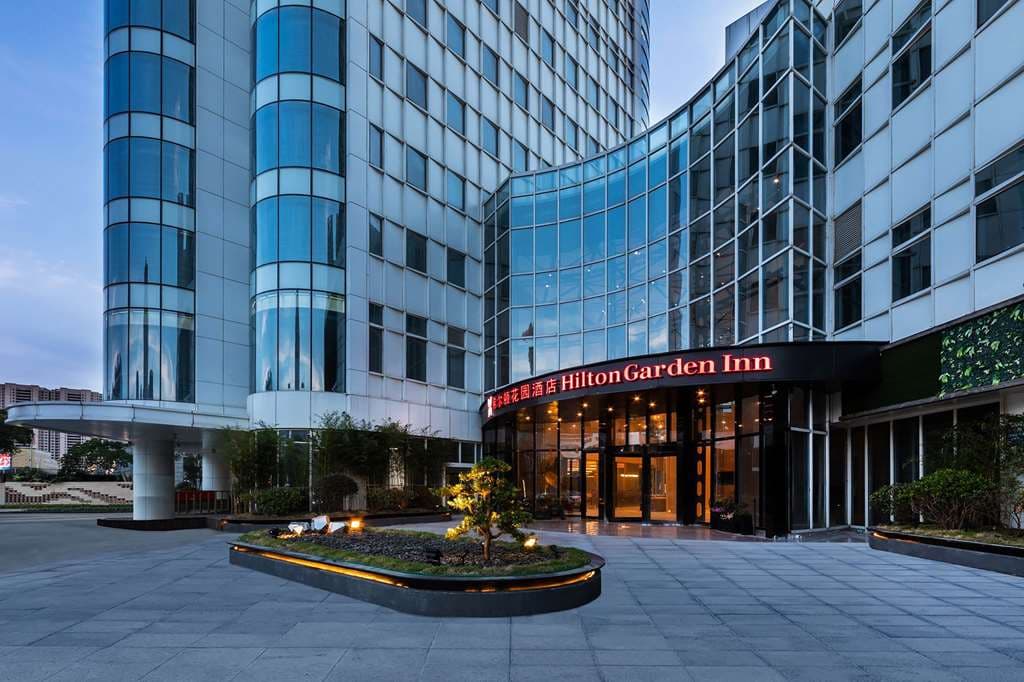 Exterior of Hilton Garden Inn Nantong Xinghu at twilight, featuring a modern glass facade with warm lobby lighting, set against the Nantong city skyline.