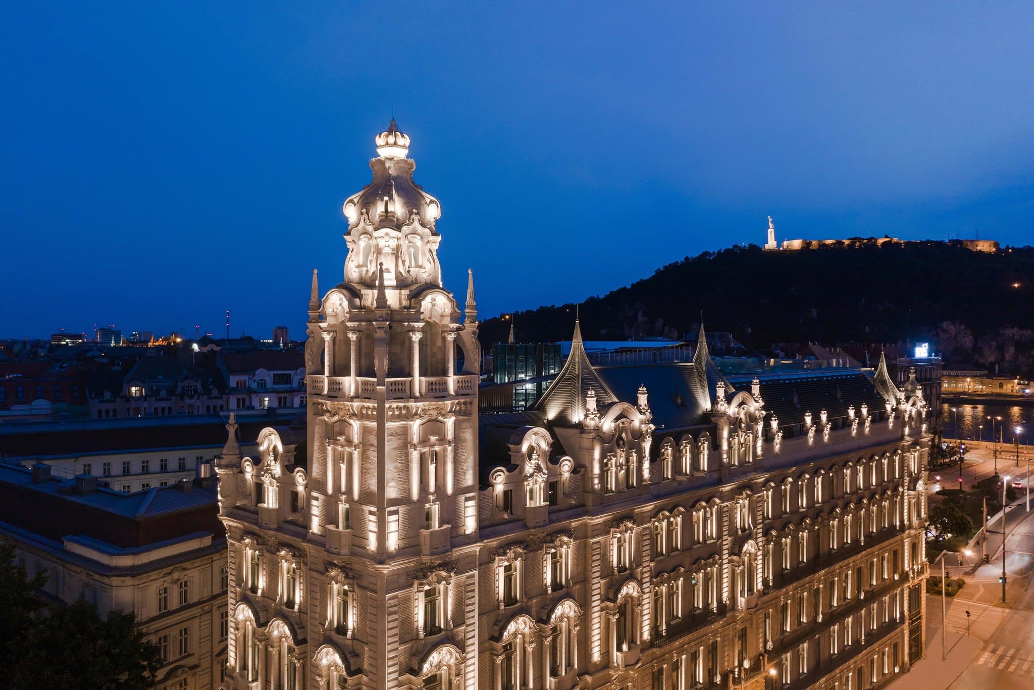 The illuminated Neo-Baroque facade of Matild Palace at night, featuring intricate stucco work and the Elizabeth Bridge in the background.